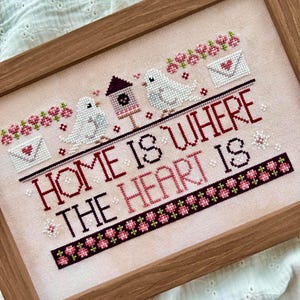 May include: Cross-stitch artwork in a wooden frame. The design features the text "HOME IS WHERE THE HEART IS" in black and burgundy thread. Two white birds, a birdhouse, and floral accents are also stitched into the piece. The background is a light pink fabric.