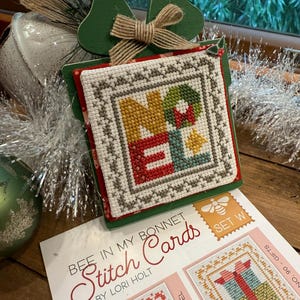 May include: A set of cross stitch patterns for Christmas cards. The patterns feature a festive red, green, and white color scheme. The patterns are titled "Bee in My Bonnet Stitch Cards" by Lori Holt.