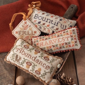 May include: A collection of fall-themed cross-stitch ornaments. The ornaments include the words "FALL", "Bountiful", "Harvest", and "Abundance". The ornaments are in various shapes and sizes, with autumnal colors and decorative stitching.
