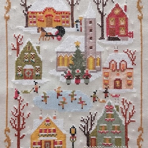 May include: A cross-stitch winter village scene with snow-covered houses, trees, and figures ice skating. The houses are various colors, and the scene is framed with a gold border. A horse-drawn carriage is also visible.