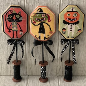 May include: Three Halloween-themed cross-stitch decorations. Each features a cartoon character: a black cat, a witch, and a pumpkin. They are mounted on black rods with wooden spools and black ribbons. The cat is on a red background, the witch on yellow, and the pumpkin on green.