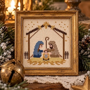 May include: A square cross-stitch embroidery in a gold frame depicting a nativity scene. The design features three figures in blue, brown, and yellow tones under a brown stable structure with a star above, all on a white fabric background.