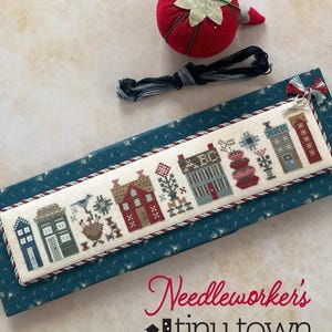 May include: A cross-stitch sampler featuring a tiny town design with houses in various colors. The sampler is on a blue fabric with a red and white rope trim. A red pin cushion with a green flower design and black thread are also in the image. The text "Needleworker's tiny town" is in the image.