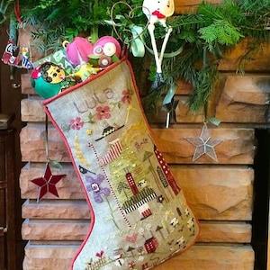 May include: A beige Christmas stocking with red trim and a colorful embroidered design. The stocking is filled with small gifts and is hanging on a brick fireplace. The stocking is personalized with the name "Lula".
