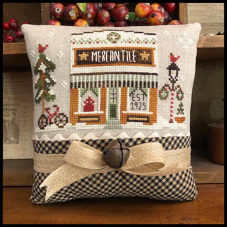 May include: A cross-stitch pillow featuring a vintage-style storefront with the text "Mercantile Est. 1925". The pillow is decorated with a brown and white checkered fabric, a burlap ribbon, and a bell.