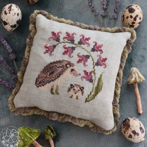 May include: A beige pillow with a brown fringe border features a cross-stitch design of two hedgehogs under a cluster of pink and purple flowers. The pillow is on a blue-green surface with quail eggs and mushrooms.