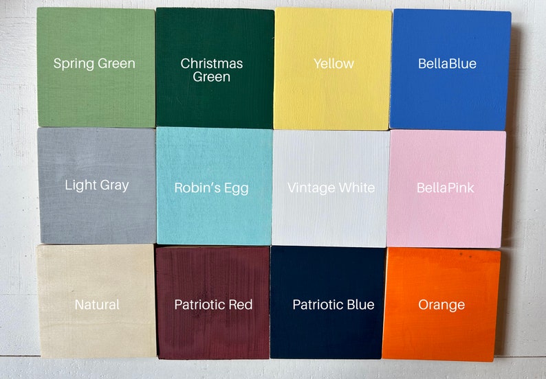 May include: Twelve paint color samples displayed on square wooden blocks. The colors are: Spring Green, Christmas Green, Yellow, BellaBlue, Light Gray, Robin's Egg, Vintage White, BellaPink, Natural, Patriotic Red, Patriotic Blue, and Orange.