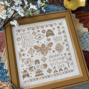 May include: A framed cross-stitch embroidery featuring a bee, honeycomb, flowers, and the alphabet in a brown and white color scheme. The embroidery is set against a beige background and framed in a brown wood frame.