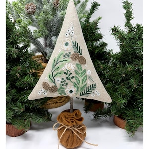 May include: A decorative, handcrafted Christmas tree with a beige fabric exterior. The tree is adorned with embroidered green leaves, white flowers, and brown pinecones. It stands on a wooden stick, secured by a brown felt base tied with twine. The background features evergreen trees.
