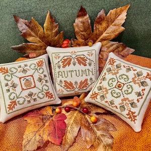 May include: Three small, square, cream-colored pillows with embroidered autumn-themed designs. The pillows are decorated with fall leaves, pumpkins, and acorns. The center pillow has the word "AUTUMN" embroidered in brown.