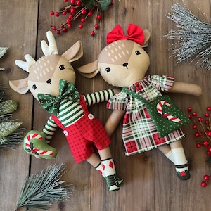 May include: Two handmade deer dolls with festive holiday attire. One doll wears red plaid shorts and a green striped shirt with a bow tie. The other doll wears a plaid dress and carries a green bag with a candy cane design.