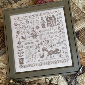 May include: A framed cross-stitch sampler featuring brown and muted-tone embroidery on a light fabric background. The design includes the alphabet in uppercase and lowercase letters, numbers 1-0, a house, a squirrel, and floral motifs.