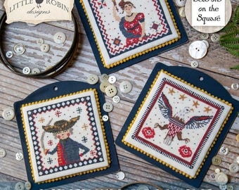May include: Three cross-stitch designs on navy blue fabric with yellow trim. The designs feature patriotic themes: a woman holding a torch, a man with a bow, and an eagle with stars and stripes. The words "LIBERTY SQUARE" are at the bottom.