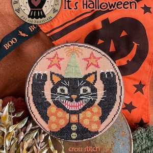 May include: A circular cross stitch piece featuring a black cat with a wide grin, wearing a light green party hat and an orange polka dot bow tie. The cat is set against a peach background with two orange stars. Text reads Yippee! It's Halloween.