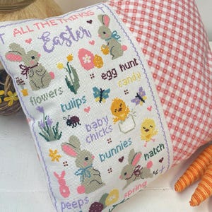 May include: White pillow with a cross-stitch design featuring Easter-themed illustrations and text. The text reads "All the things Easter" and includes words like "flowers", "tulips", "baby chicks", "bunnies", "peeps", "spring", "egg hunt", and "hatch".