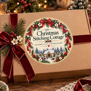 May include: A rectangular brown cardboard box wrapped with a deep red velvet ribbon and a decorative sprig of pine needles, red berries, and white flowers. A circular label features the text The Christmas Stitching Cottage Anabella's Needleart.