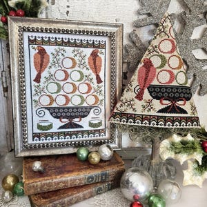 May include: A framed cross-stitch featuring two birds and a decorative bowl with eggs. A matching tree-shaped decoration stands beside it. The scene is decorated with vintage books and Christmas ornaments in silver, green, and gold.
