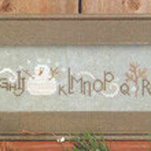 May include: A framed cross-stitch sampler with a blue background and white snowmen. The sampler features the alphabet in a whimsical font with the letters "A" through "Z" and the words "Deck the Halls".