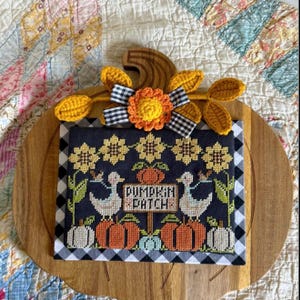 May include: A rectangular cross-stitch wall hanging on a dark blue background featuring two white geese, orange pumpkins, and yellow sunflowers. The text PUMPKIN PATCH is centered. It is topped with a crocheted sunflower and a black and white gingham bow.