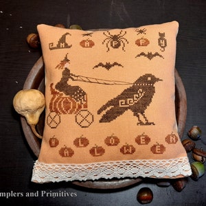 May include: A hand-stitched pillow with a whimsical Halloween design featuring a witch riding a pumpkin carriage pulled by a crow. The pillow has a peach background with black stitching and a lace trim.