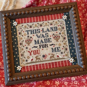 May include: Framed cross-stitch artwork with the text "THIS LAND WAS MADE FOR YOU and ME." The piece features a wooden frame with a beaded edge, red and blue accents, and small white flower embellishments. The artwork has a patriotic theme.