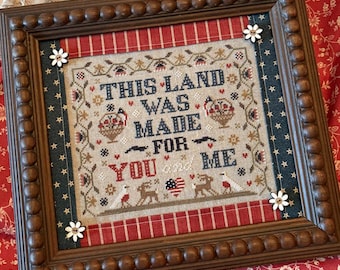 May include: A framed cross-stitch piece with the text "THIS LAND WAS MADE FOR YOU and ME." The wooden frame has a beaded edge, with red and blue accents, and small white flower embellishments. The artwork has a patriotic theme, with a rustic aesthetic.
