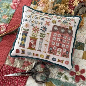 May include: A white decorative pillow with a cross-stitch design featuring a red house, spools, and the text "button house". The pillow has a teal border and is placed on a floral quilt. A pair of ornate scissors is in the foreground.