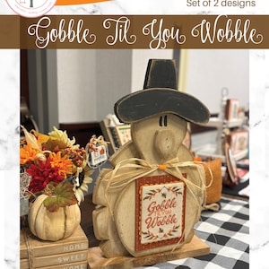 May include: Wooden Thanksgiving decoration featuring a turkey wearing a black hat and a tag that says "Gobble Til You Wobble." The turkey is made of light-colored wood and has a rustic, farmhouse style. A fall-themed floral arrangement is in the background.