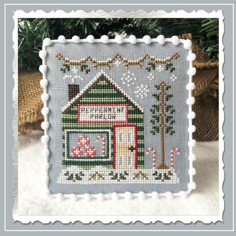 Puede incluir: Un patr&oacute;n de punto de cruz de una peque&ntilde;a casa de madera verde con un letrero que dice "Peppermint Parlor". La casa est&aacute; decorada con bastones de caramelo rojos y blancos y tiene una ventana con bastones de caramelo rojos y blancos en el interior. La casa est&aacute; rodeada de nieve y un peque&ntilde;o &aacute;rbol de hoja perenne.
