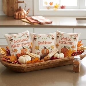 Könnte beinhalten: Drei quadratische Dekokissen mit Kreuzstichmotiven von Kürbissen, Eicheln und Herbstblättern. Die Kissen tragen die Aufschriften "Autumn Blessings", "Gather" und "Hello Autumn". Sie sind in einer hölzernen Teigschale arrangiert.
