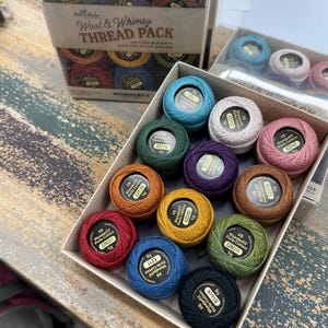 May include: A collection of WonderFil Perle Cotton thread spools in a cardboard box, featuring various colors including red, blue, green, yellow, orange, purple, and black. Each spool is labeled with its color name, weight, and product code.