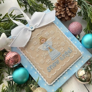 May include: A white and blue cross-stitch ornament with an angel design and the text "Snow Angel". The ornament is on a blue background with white stars.