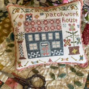 May include: A small, square, embroidered pillow with the words "patchwork house" stitched on it. The pillow features a cross-stitched house design in blue, red, and green. The pillow is trimmed with a pink, fringed edge. A pair of decorative scissors rests on a quilted surface.