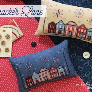 May include: Two decorative pillows with cross-stitch house designs, one cream and one blue. The cream pillow has red, white, and blue accents. The blue pillow has white and red accents. The image also includes buttons and the text "Firecracker Lane".