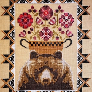 May include: Cross-stitch embroidery of a brown bear with a pot of purple flowers on its head. The bear is surrounded by a black and white geometric border.
