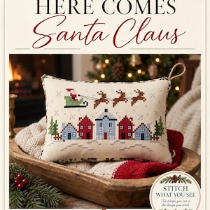 May include: A rectangular cross-stitch cushion featuring a winter village scene with red and blue houses, green pine trees, and Santa Claus in a sleigh pulled by three reindeer. The text reads "ANABELLA'S NEEDLEART" and "HERE COMES Santa Claus".