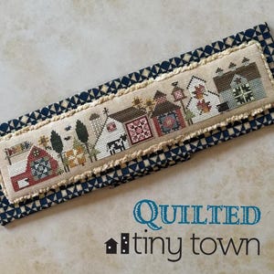 May include: A cross-stitch piece featuring a quaint village scene with various buildings, trees, and a cow. The piece is framed by a blue and white patterned fabric. The text "Quilted tiny town" is displayed in the lower right corner.