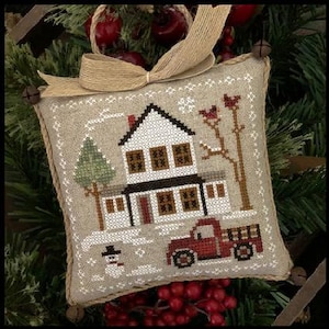 Op de afbeelding: Een kruissteek ornament met een wit huis met rode versiering, een rode vrachtwagen en een sneeuwpop. Het huis heeft bruine ramen en een bruine deur. De vrachtwagen heeft een rode laadbak en een witte cabine. De sneeuwpop draagt een zwarte hoed en een zwarte sjaal. Het ornament is omgeven door een bruin jute lint.