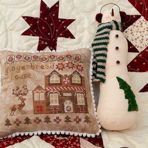 May include: A brown and tan embroidered pillow with the text "Gingerbread House" and a small embroidered image of a gingerbread house, a reindeer, and trees. The pillow is on a white and red quilted fabric with a large red star in the background. A white snowman with a green and brown striped scarf and red earmuffs is to the right of the pillow.