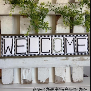 May include: A framed sign with the word "WELCOME" in large, white, block letters. The letters are outlined in black and set against a black and white checkered background. The letter "O" is decorated with a green wreath. The sign is set on a white, rustic wooden base.