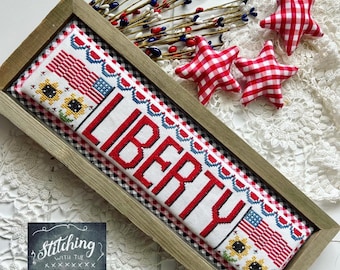 May include: A framed cross-stitch piece featuring the word "LIBERTY" in red lettering on a white background. The piece is bordered with a red and white gingham trim. The frame is made of wood and has a rustic finish. The image also includes a sign that reads "Stitching with the Housewives".