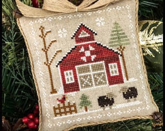 May include: A cross-stitch ornament depicting a red barn with a white star and a white fence. The barn is surrounded by trees and snow. There are three black sheep in front of the barn.