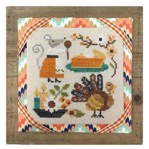 May include: Cross-stitch artwork featuring Thanksgiving-themed elements. A turkey, pumpkin pie, and a mouse are depicted in the cross-stitch design. The artwork is framed in a wooden frame with a patterned fabric border and a decorative ribbon.