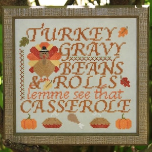 May include: Cross-stitch artwork in a wooden frame. The design features the words "TURKEY GRAVY BEANS & ROLLS" and "lemme see that CASSEROLE" in orange and brown. A turkey, pumpkins, and food illustrations complete the Thanksgiving theme.