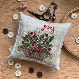 May include: A square, off-white embroidered pillow with the word "JOY" in red. The pillow features a Christmas basket design with holly, berries, and a candy cane. Small white buttons and a pair of black scissors are scattered around the pillow on a wooden surface.