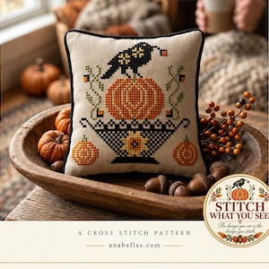 May include: A square beige fabric pillow featuring a cross-stitch design of a black crow perched on an orange pumpkin, set within a black basket. The pattern includes green vines, small orange berries, and two additional orange pumpkins.
