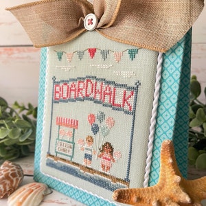 May include: A cross-stitch embroidery of a boardwalk scene with a cotton candy stand, balloons, and two children. The embroidery is framed in a light blue frame with a white rope border and a turquoise background. The frame is decorated with a brown burlap bow.