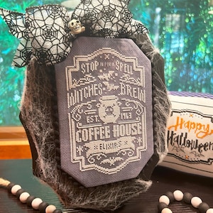 May include: A black framed cross-stitch with a purple background and white embroidery. The embroidery reads "Stop in for a Spell Witches Brew Est. 1713 Coffee House Elixir's". A black and white spiderweb bow with a skull charm is on top of the frame. A white pillow with black embroidery reads "Happy Halloween".