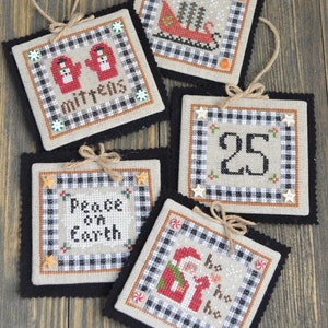 May include: A collection of five square, cross-stitched Christmas ornaments. Each features a different holiday design, including mittens, a sleigh, the number 25, Santa Claus, and the phrase "Peace on Earth." They have a black and white checkered border and are trimmed with black felt.
