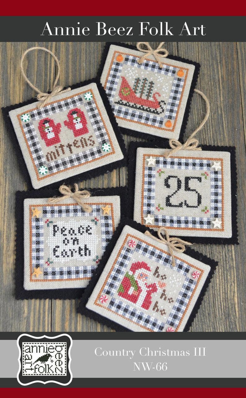May include: A collection of five square, cross-stitched Christmas ornaments. Each features a different holiday design, including mittens, a sleigh, the number 25, Santa Claus, and the phrase "Peace on Earth." They have a black and white checkered border and are trimmed with black felt.